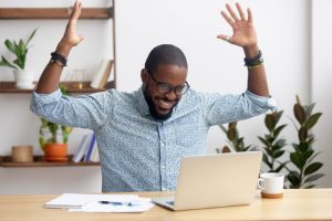 Professional man cheering with his hands up while looking at a computer as he updates his executive resume